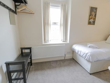 A bedroom with a bed covered in white linens a window with curtains a black bench with hangers on the wall and framed artwork at Gwelfor in Morfa Bychan