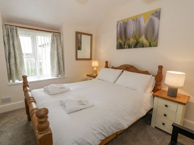 A bedroom with a wooden bed with white linens two bedside tables with lamps a window with curtains and a wall mirror at Gwelfor in Morfa Bychan
