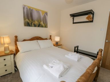 A bedroom with a wooden bed white bedding bedside tables with lamps a wall shelf with hangers and a picture of white flowers at Gwelfor in Morfa Bychan