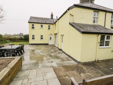A paved patio area with outdoor dining furniture next to a yellow two-story house at Gwelfor in Morfa Bychan