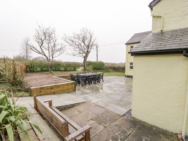 An outdoor patio area with a large wooden table and chairs near a yellow house with tiled roof at Gwelfor in Morfa Bychan