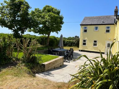 A garden with outdoor dining table and chairs, trees, grass, and a yellow house at Gwelfor in Morfa Bychan