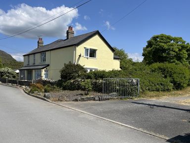 A yellow two-story house with chimney stacks by a roadside with bushes and a metal gate at Gwelfor in Morfa Bychan