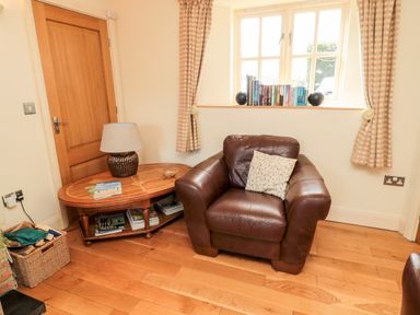 A room corner with a brown leather armchair a wooden side table with a lamp books on it and a window with books and curtains at The Granary Longframlington near Alnwick