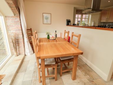 A dining area with a wooden table and six wooden chairs near a window and an open kitchen at The Granary in Longframlington near Alnwick