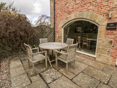 An outdoor patio area with a round wooden table and four wooden chairs beside a brick building with a large arched window at The Granary in Longframlington near Alnwick