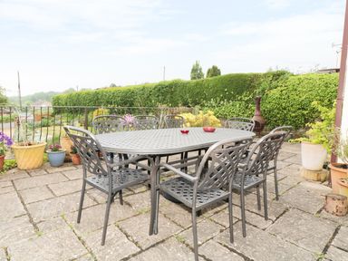 An outdoor patio area with a metal table and eight matching chairs surrounded by potted plants and greenery at Lynton House Ruardean Hill near Drybrook