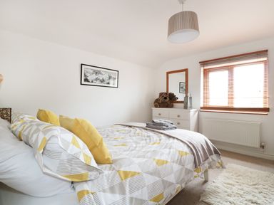 A bedroom with a bed dressed with patterned bedding and yellow cushions a white dresser with a mirror and a window with wooden blinds at Lynton House Ruardean Hill near Drybrook