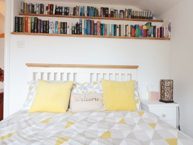 A bed with yellow pillows and a welcome cushion under two wooden shelves filled with books at Lynton House in Ruardean Hill near Drybrook