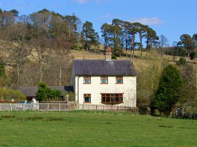 A detached two-story house with a fenced yard in front of trees and grassy hills at Hartam House in Falstone near Kielder and Bellingham