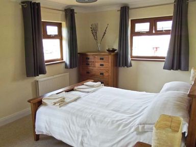 A bedroom with a wooden bed white bedding folded towels a wooden chest of drawers two windows with dark curtains and a small lamp on bedside table at Hartam House in Falstone near Kielder and Bellingham