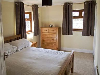 A bedroom with a wooden bed a chest of drawers a bedside table and two windows with curtains at Hartam House in Falstone near Kielder and Bellingham