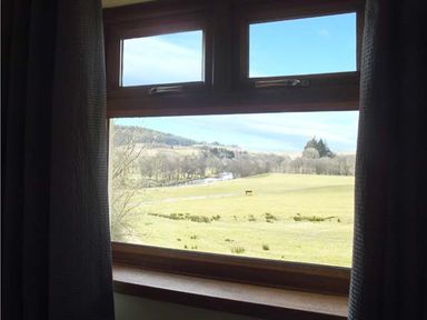 A view of a grassy field and distant hills through a wooden window with curtains at Hartam House in Falstone near Kielder and Bellingham