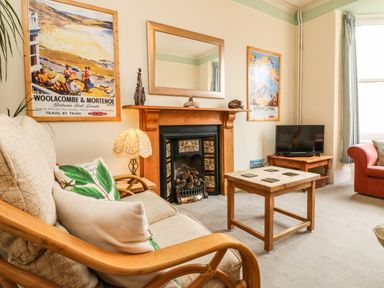 A living room with a beige armchair and pillows a wooden coffee table a fireplace with a mirror above and a small television on a wooden stand at Penryn in Ilfracombe