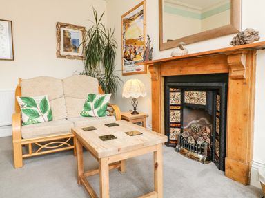A sitting area with a wooden framed sofa with cushions a wooden coffee table a side table with a lamp a fireplace with decorative tiles and pinecones a tall plant and framed pictures on the walls at Penryn in Ilfracombe