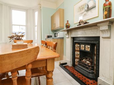 A dining area with wooden table and chairs a decorative fireplace and kitchen cabinets at Penryn in Ilfracombe