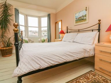 A bedroom with a bed with checkered bedding two bedside tables with lamps a large mirror a painting a window with blue curtains and two chairs by the window at Penryn in Ilfracombe
