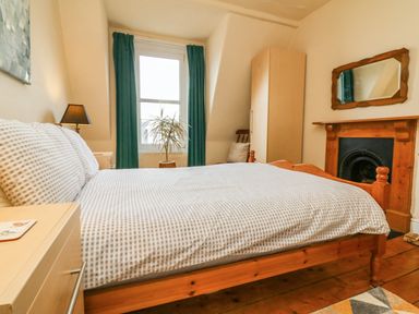 A bedroom with a wooden bed a bedside table with a lamp a window with green curtains a plant a wardrobe a chair a mirror above a wooden fireplace and wooden floorboards at Penryn in Ilfracombe