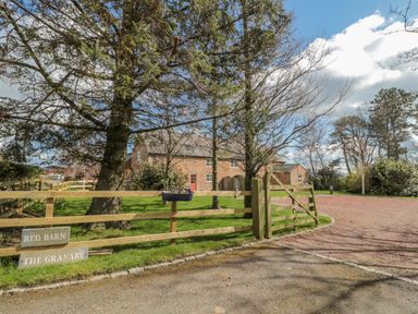 A brick house behind a wooden fence and gate with trees and a gravel driveway at The Red Barn in Longframlington near Alnwick