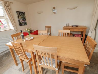 A dining room with a wooden table and six chairs a bottle of wine and a plate with bread on the table a wooden sideboard with a bowl salt and pepper shakers on the sideboard a window with curtains and a picture on the wall at The Red Barn in Longframlington near Alnwick