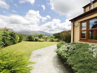 A garden with bushes, a lawn, and a paved pathway beside a house with mountains in the background at Ddol Helyg Farmhouse in Llanrug
