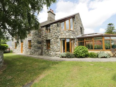A stone house with wooden framed windows and a conservatory surrounded by a lawn and plants at Ddol Helyg Farmhouse in Llanrug