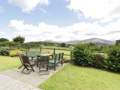 An outdoor patio with wooden chairs and benches overlooking a fenced grassy field and hills at Ddol Helyg Farmhouse Llanrug