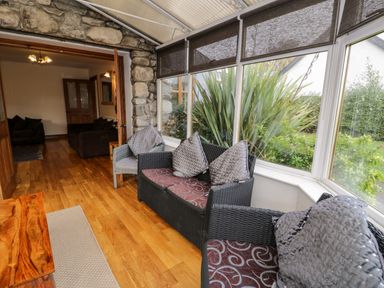 A sunroom with wicker seating and cushions overlooking a garden at Ddol Helyg Farmhouse Llanrug