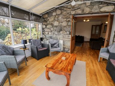 A sunroom with wicker chairs and a wooden table next to a stone wall at Ddol Helyg Farmhouse in Llanrug