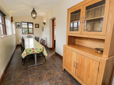 A dining room with a long table covered with a fruit-patterned tablecloth surrounded by chairs and a wooden cabinet with glass doors at Ddol Helyg Farmhouse in Llanrug