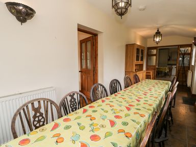 A dining room with a long table covered with a fruit patterned tablecloth and wooden chairs at Ddol Helyg Farmhouse in Llanrug