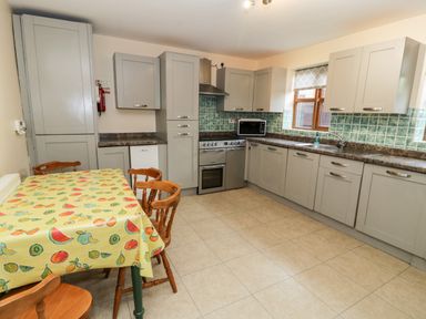 A kitchen with grey cabinets green tiled backsplash a stove microwave and wooden chairs around a table with a fruit patterned tablecloth at Ddol Helyg Farmhouse in Llanrug