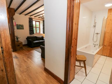 A hallway leads to a living room with sofas and a window and a bathroom with a bathtub and a wooden chair at Ddol Helyg Farmhouse in Llanrug