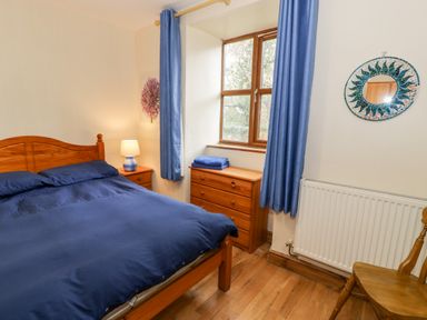 A bedroom with a bed covered in blue bedding next to wooden furniture and a window with blue curtains at Ddol Helyg Farmhouse Llanrug