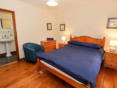 A bedroom with a wooden bed and blue bedding two wooden nightstands with lamps a blue chair and a wooden dresser at Ddol Helyg Farmhouse in Llanrug