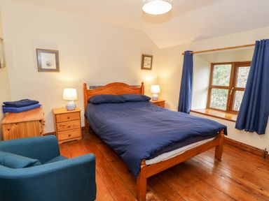 A bedroom with a wooden double bed with blue bedding two wooden nightstands with lamps a blue armchair wooden flooring and a window with blue curtains at Ddol Helyg Farmhouse in Llanrug