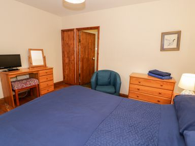 A bedroom with a bed covered with blue bedding a wooden dresser a blue armchair and a wooden desk with a TV and mirror at Ddol Helyg Farmhouse in Llanrug