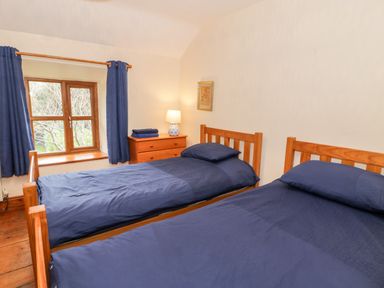 A bedroom with two single beds with blue bedding a wooden dresser with a lamp and towels and a window with blue curtains at Ddol Helyg Farmhouse Llanrug
