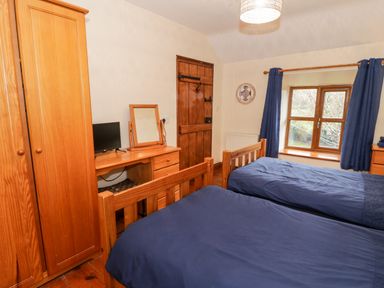 A bedroom with two single beds a wooden wardrobe desk with a mirror and a small tv and window with blue curtains at Ddol Helyg Farmhouse in Llanrug