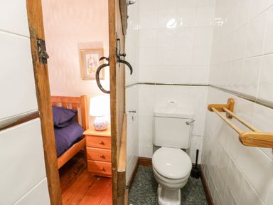 A small bathroom with a toilet and wooden handrails next to a bedroom with wooden furniture and a lamp at Ddol Helyg Farmhouse Llanrug
