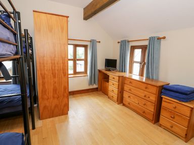 A bedroom with bunk beds a wardrobe a desk with a small TV two windows with curtains and wooden drawers at Ddol Helyg Farmhouse Llanrug