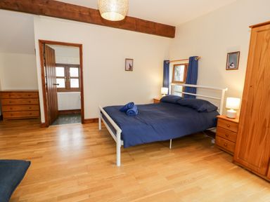 A bedroom with a double bed and blue bedding two wooden bedside tables with lamps a wooden wardrobe and dresser a door leading to a bathroom with windows and wooden accents at Ddol Helyg Farmhouse in Llanrug