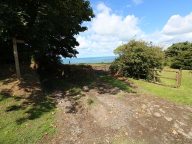 A dirt path leading to a metal gate with trees and bushes on the sides and a view of the sea at Morfa Isaf Cottage in Llangrannog