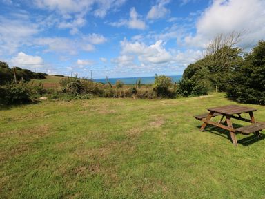 A grassy field with a wooden picnic table and bushes with a view of the sea at Morfa Isaf Cottage in Llangrannog