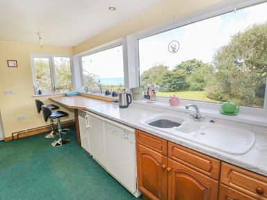 A kitchen with a countertop sink and two bar stools by a corner wooden table near large windows showing a garden and sea view at Morfa Isaf Cottage in Llangrannog