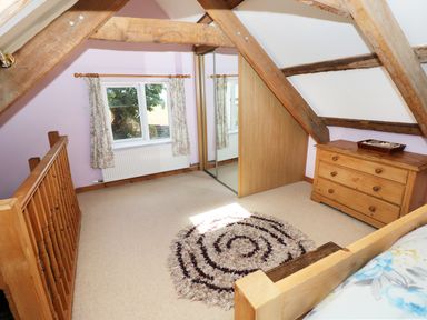An attic bedroom with wooden beams a chest of drawers a mirrored wardrobe a window with floral curtains and a circular rug on the floor at Morfa Isaf Cottage in Llangrannog
