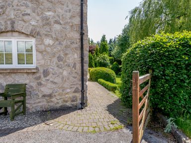 An outdoor garden area with a stone wall house a gate and a paved path surrounded by trimmed bushes at Distyll Llanfwrog near Ruthin