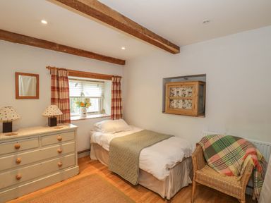 A bedroom with a single bed a wooden dresser with two lamps a wicker chair with a blanket and a small window with curtains at Pembridge Cottage in Welsh Newton