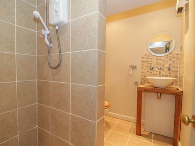 A bathroom with a tiled shower, a wooden sink stand with a round mirror and a toilet at Pembridge Cottage in Welsh Newton