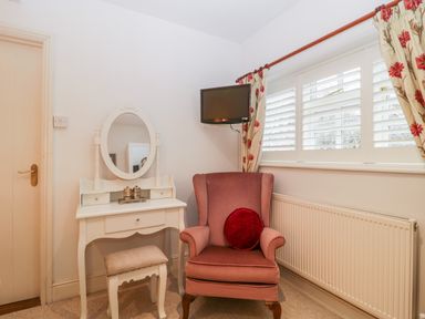 A corner with a white dressing table with mirror and stool a mauve armchair with a red cushion under a window with white shutters and floral curtains at Colly Cottage in Dottery